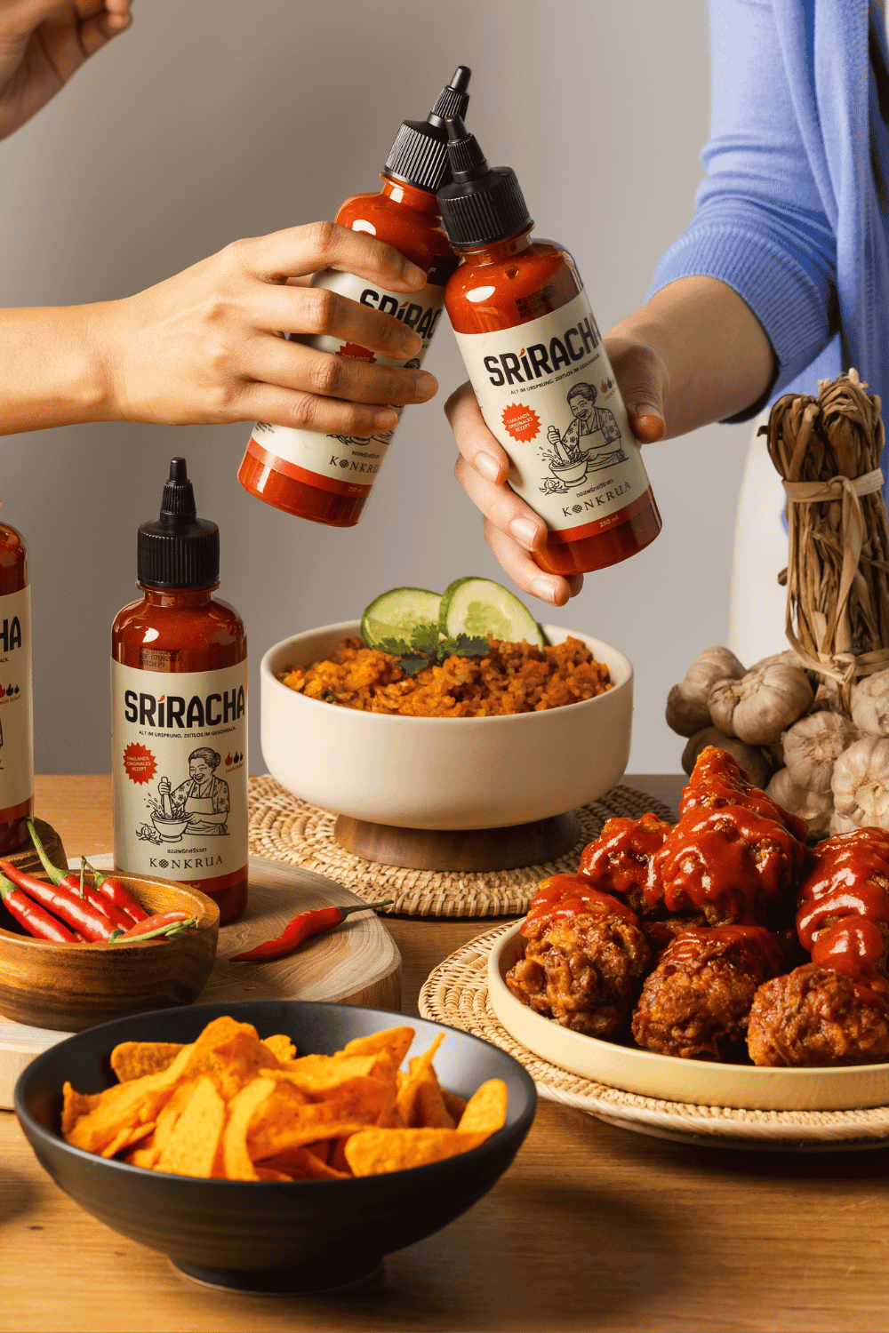 Zwei Personen halten Flaschen KONKRUA Sriracha Sauce über einen gedeckten Tisch mit gebratenem Reis, Chicken Wings und Nachos