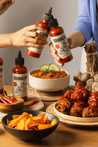 Thumbnail for Zwei Personen halten Flaschen KONKRUA Sriracha Sauce über einen gedeckten Tisch mit gebratenem Reis, Chicken Wings und Nachos