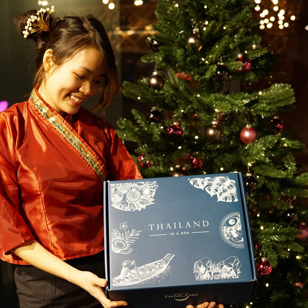 Pim, die Gründerin von KONKRUA in einer Thai Kostüm, hält Thailand in a box in der Hand und steht vor dem Weihnachtsbaum. Hier kann man Geschenkset von KONKRUA kaufen.